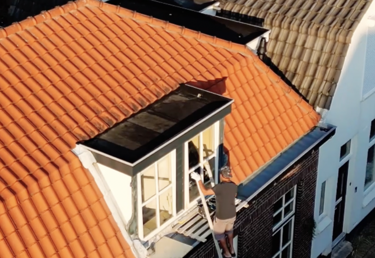 Schilder aan het werk aan een kozijn op het dak van een woning met oranje pannendaken